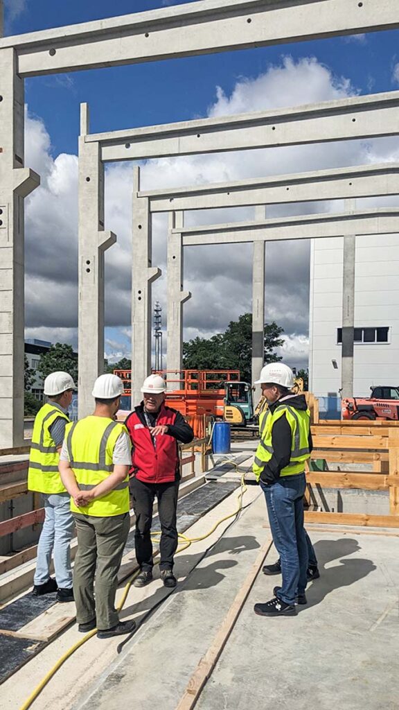 Gruppe von Menschen mit Bauhelmen und Warnwesten auf der Baustelle. Eine Person erklärt zusammenhänge auf der Baustelle.
