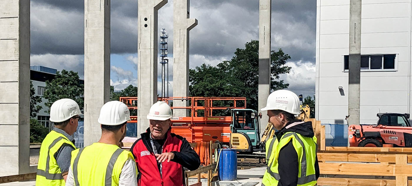 Gruppe von Menschen mit Bauhelmen und Warnwesten auf der Baustelle. Eine Person erklärt zusammenhänge auf der Baustelle.